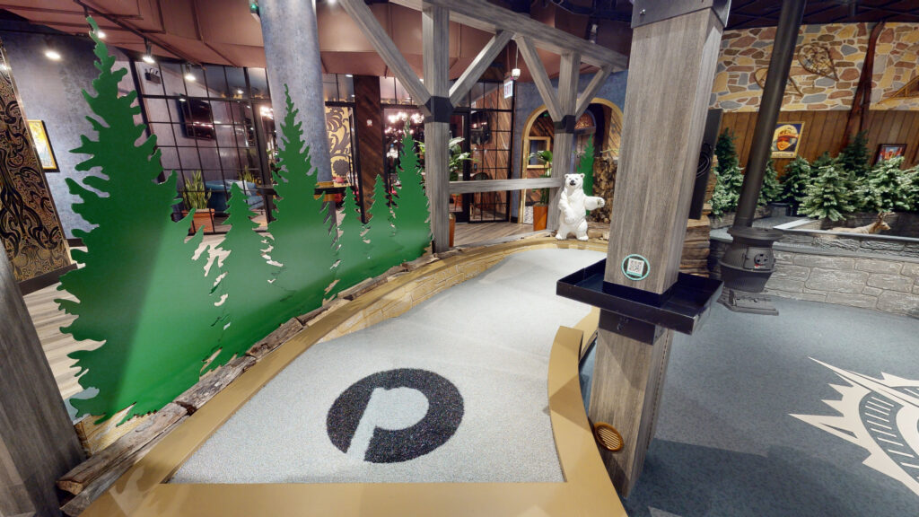 Indoor mini-golf area with green tree silhouettes along a wooden barrier and a white polar bear statue by a pillar in a rustic setting.