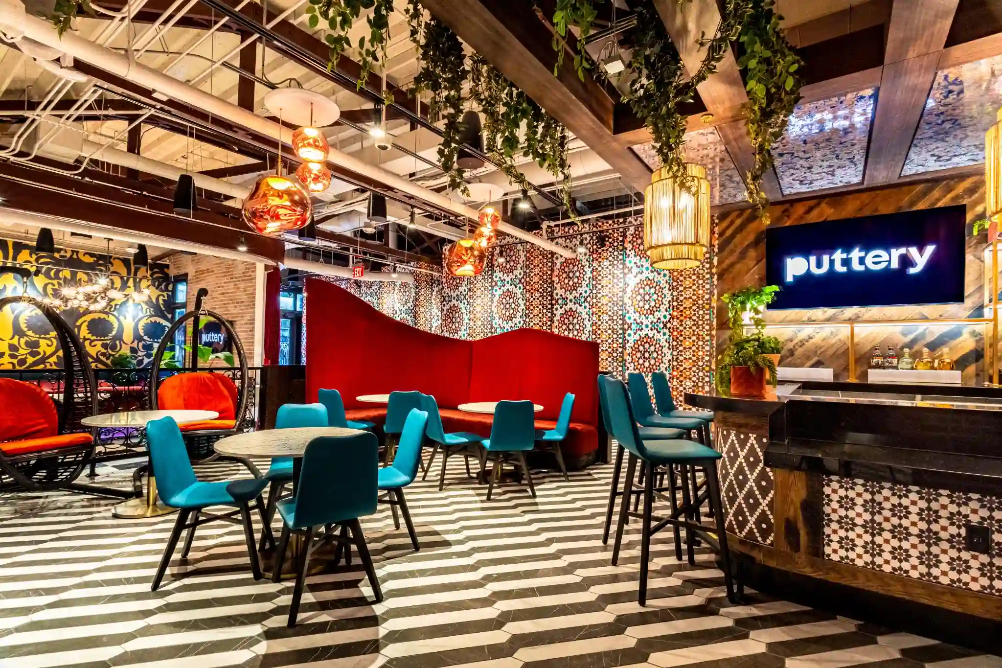 Banquette and bar area at Puttery Miami with teal chairs, a red curved banquette, striped floors and statement lighting.