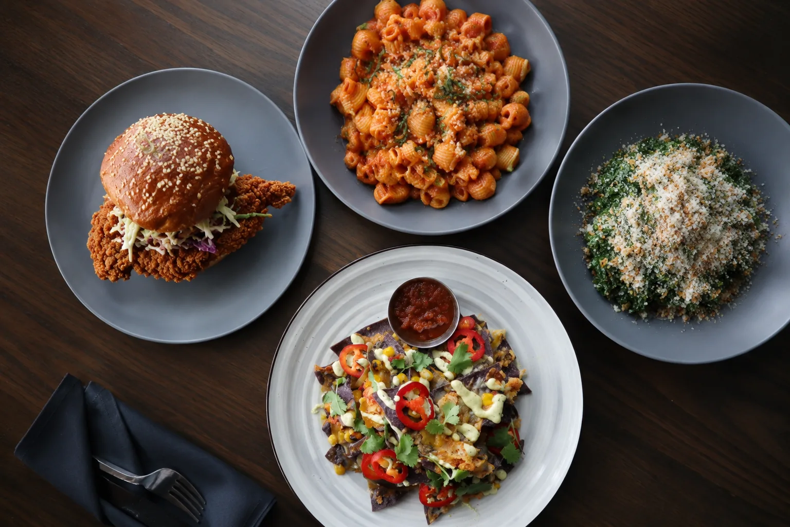 Overhead view of a Puttery New York City food spread with a crispy chicken sandwich, pasta, nachos and a greens dish.