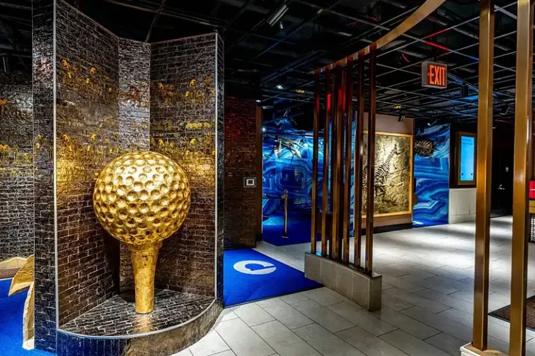 Giant gold golf ball on a tee beside Egyptian hieroglyph walls and a dinosaur fossil wall at the History Museum course entrance at Puttery New York City