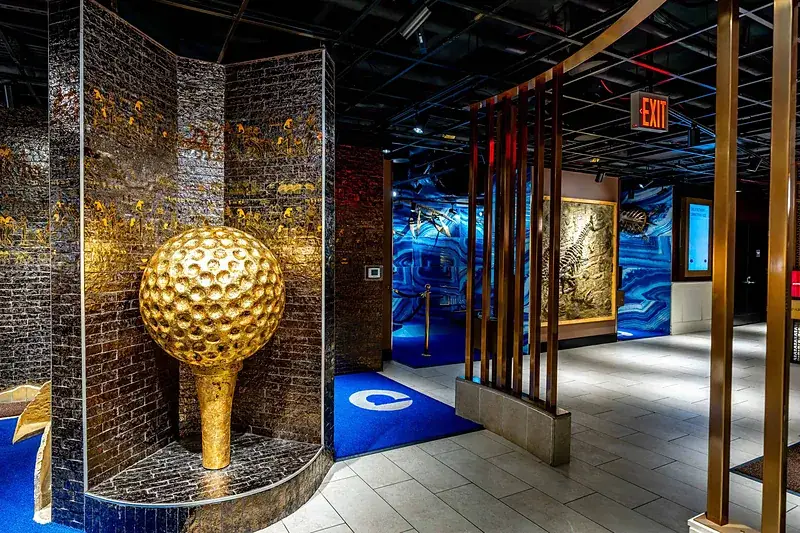 Giant gold golf ball on a tee beside Egyptian hieroglyph walls and a dinosaur fossil wall at the History Museum course entrance at Puttery New York City