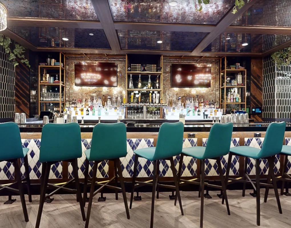 Full-service bar at Puttery Minneapolis with teal stools, two TVs, patterned tile and illuminated shelving.