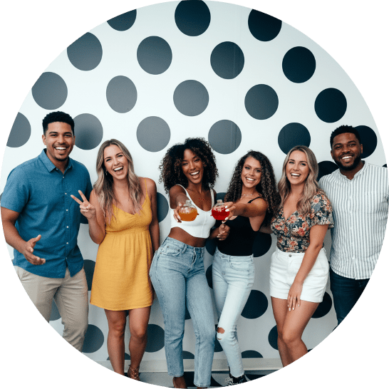 Six diverse friends posing together, toasting with cocktails in front of a polka-dot backdrop on a party night.