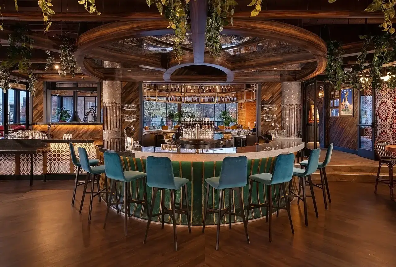 Circular bar with teal upholstered stools around a marble counter, warm lighting, and hanging greenery in a stylish lounge.