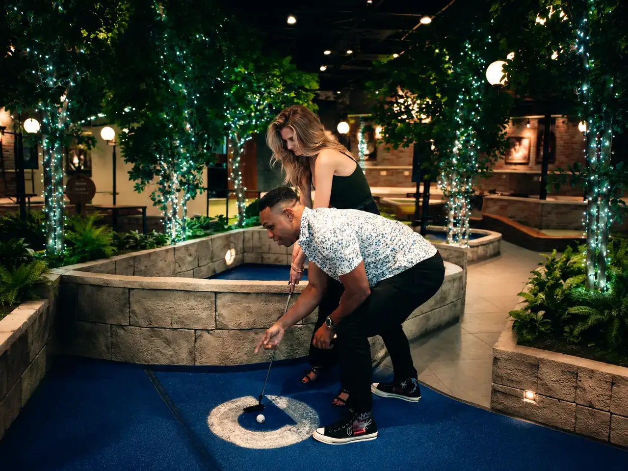 Couple lining up a putt together on a blue indoor mini golf course surrounded by illuminated trees at Puttery.