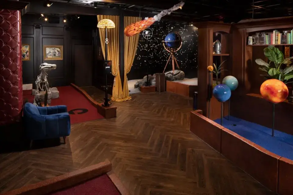 Puttery Dallas (The Colony) Library Course featuring a dinosaur skeleton, planets, a telescope, bookshelves, and a meteor suspended above the indoor mini golf course.