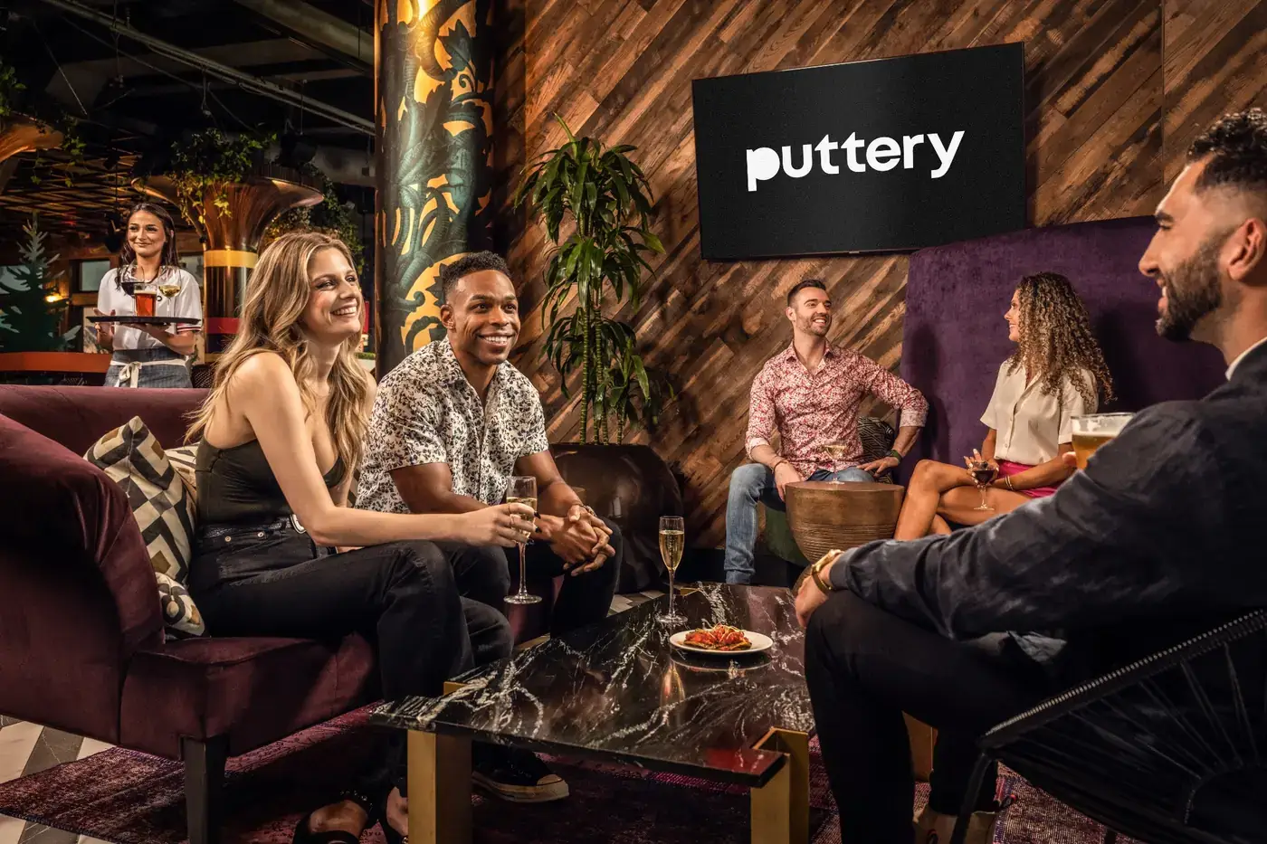 Friends socializing with cocktails and small bites in a stylish lounge at Puttery while a server approaches with drinks.