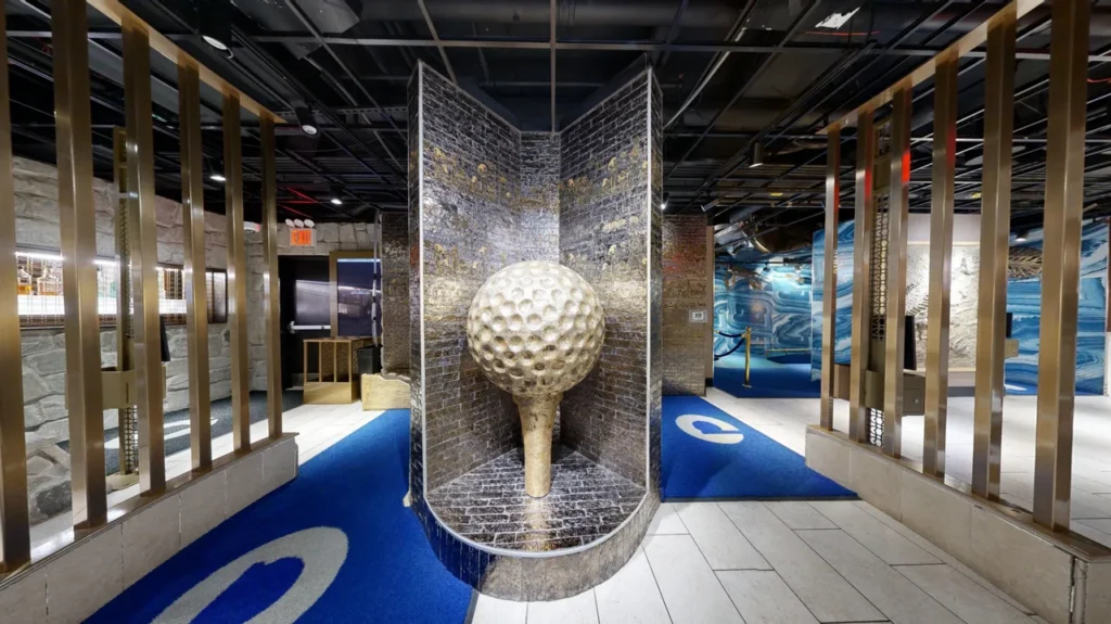 Indoor mini golf course at Puttery NYC with a large golf-ball sculpture, blue putting lanes and metallic architectural details.
