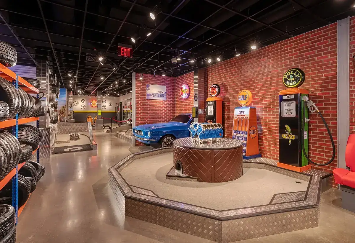 Auto shop-themed mini golf hole at Puttery Pittsburgh featuring vintage gas pumps, a blue classic car, stacked tires, and exposed brick walls.