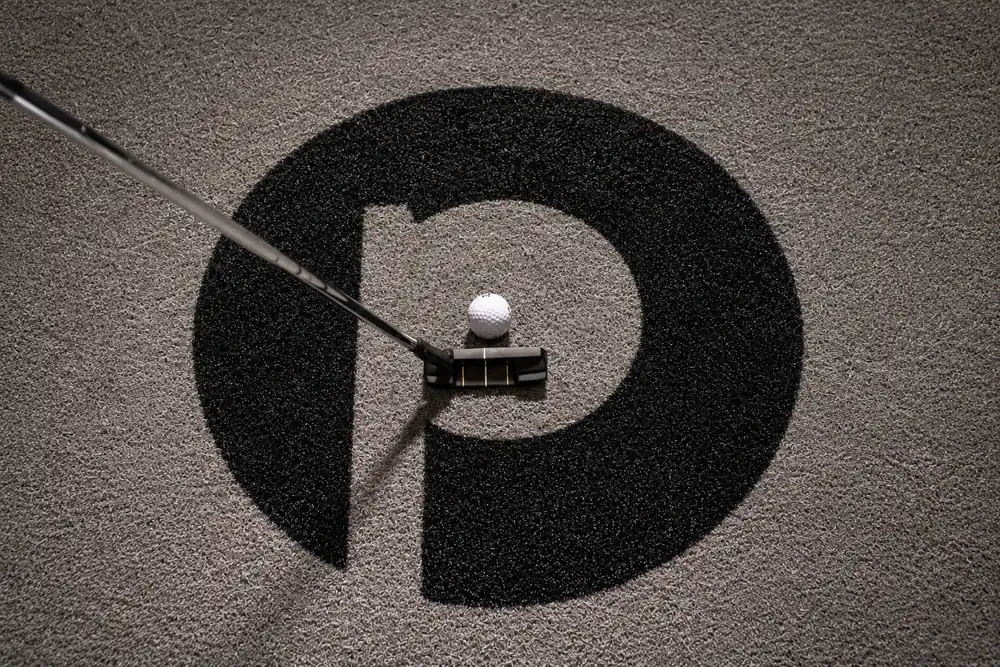 Overhead view of a putter and white golf ball positioned over the black Puttery logo on gray indoor mini golf turf.