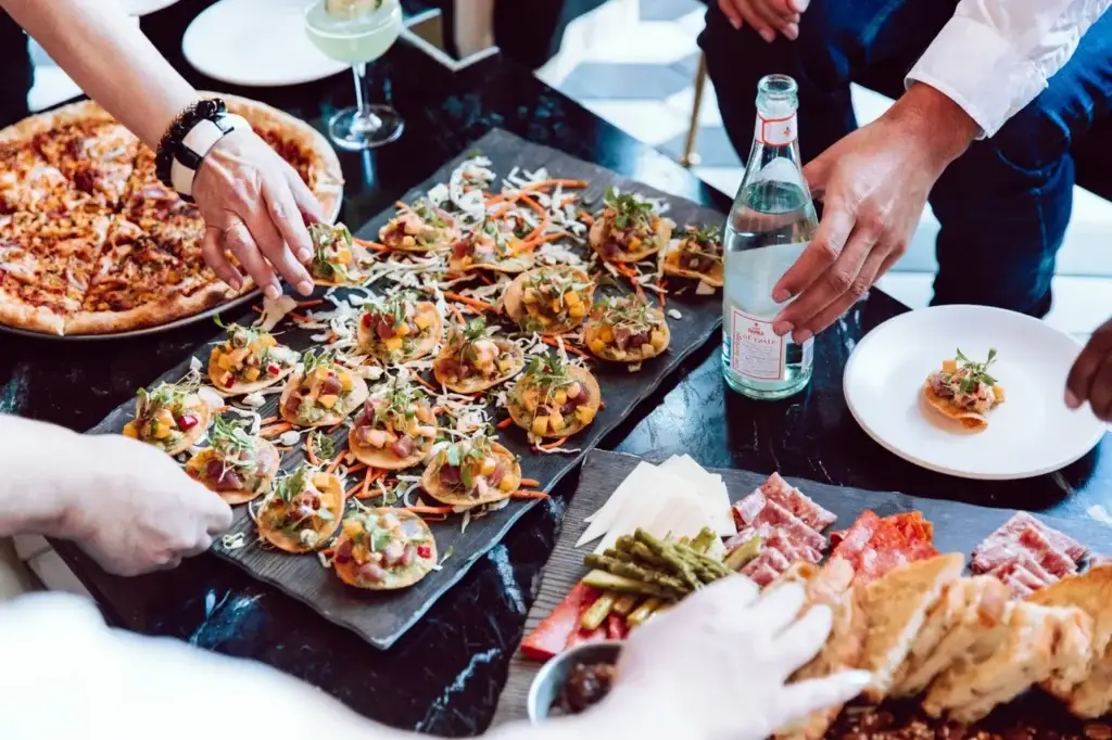Guests serving themselves from a catering spread with small bites, charcuterie items, pizza and drinks at a corporate event.