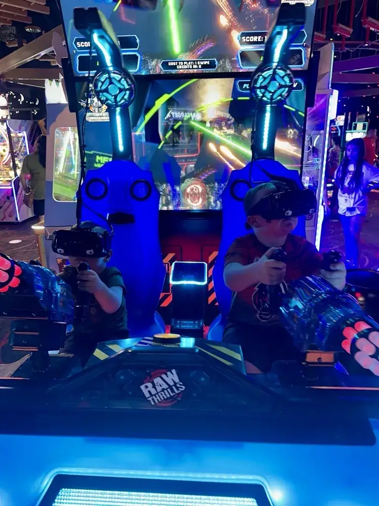 Two young kids wearing VR headsets at an arcade game marked cost to play one swipe, credits in zero