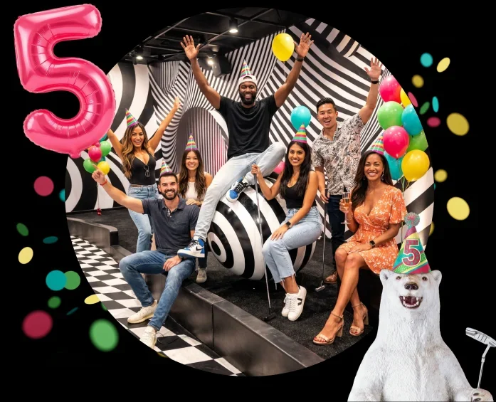Diverse group posing for a 5th birthday celebration in a black-and-white swirl party room, with party hats, balloons, confetti, a pink '5' balloon, and a white polar bear figure nearby.