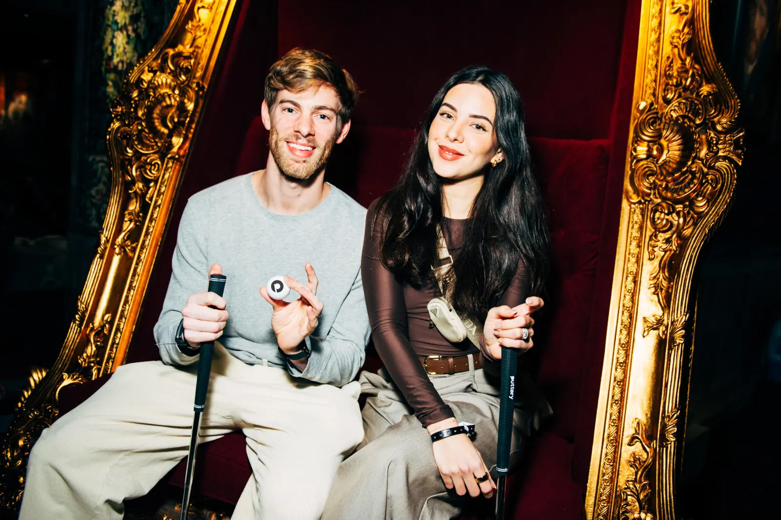 Couple seated on an ornate red throne-style chair holding putters inside Puttery.
