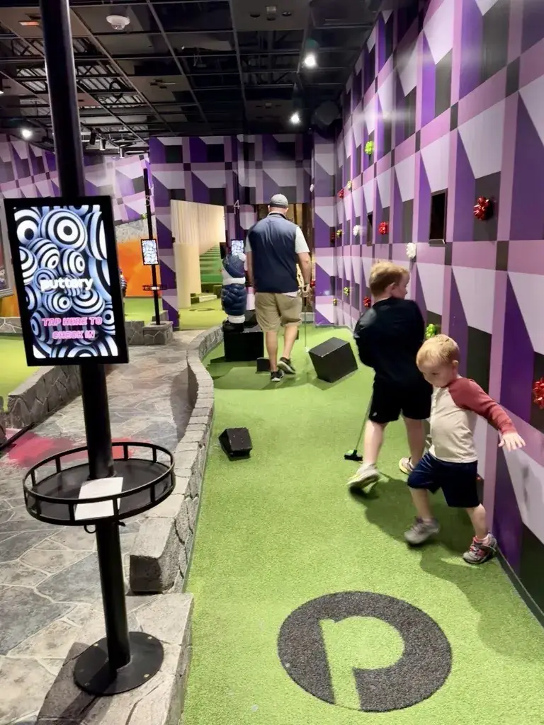 Two kids walking a colorful indoor mini golf course at Puttery past a tap to check in scorecard kiosk