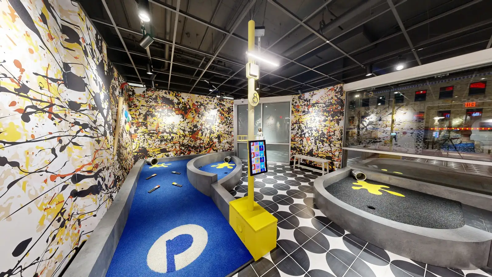 Splatter art-themed mini golf hole at Puttery Kansas City with bold paint-splashed walls, blue and black putting surfaces and graphic floor tiles.