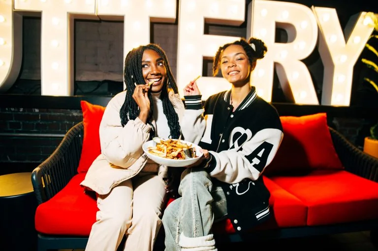 Two friends sit on a red couch, sharing fries from a plate as one eats and the other smiles, with big illuminated letters behind them.