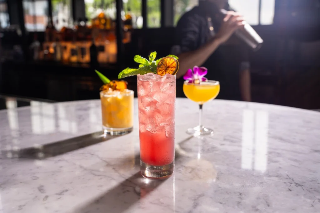 Three colorful craft cocktails displayed on a marble bar while a bartender prepares a drink behind them.