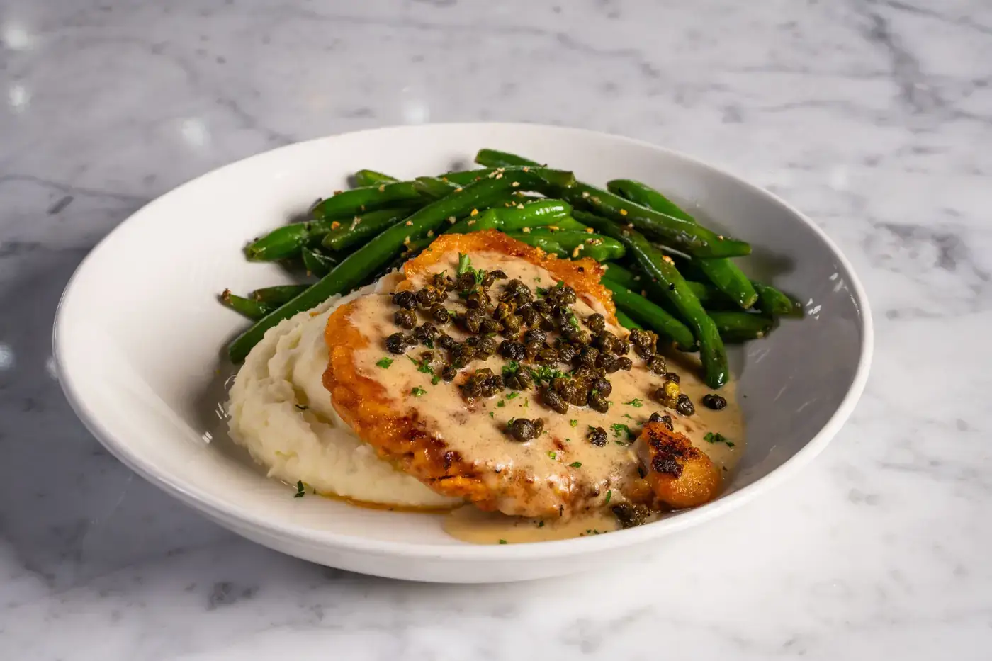 Chicken Piccata at Puttery featuring seared chicken over creamy garlic mashed potatoes, topped with lemon caper cream, fried capers and parsley, with sautéed green beans served alongside.