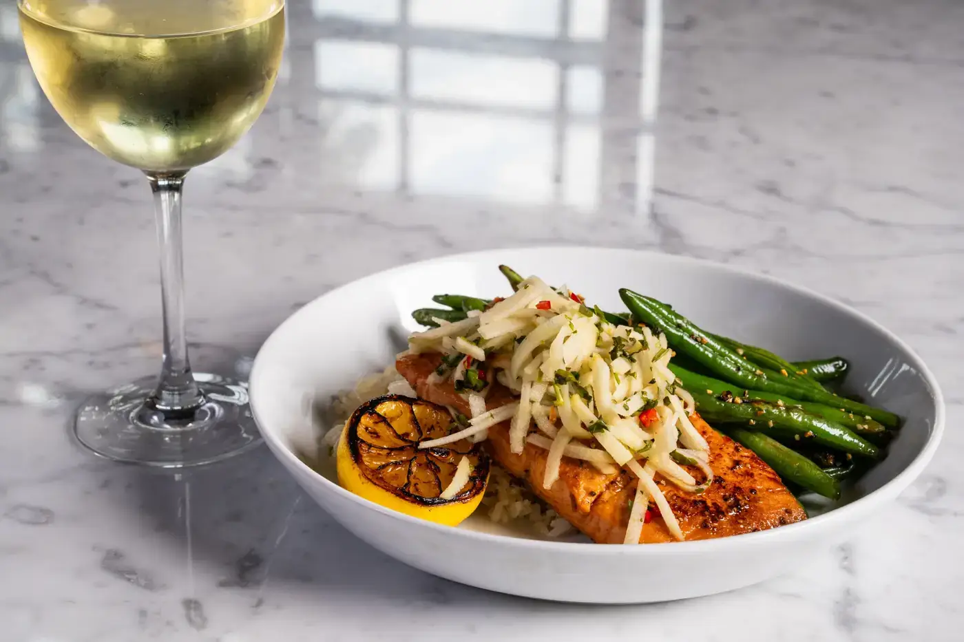 Miso Salmon at Puttery with marinated salmon served over ginger rice and topped with fresh apple slaw, accompanied by sautéed green beans, a charred lemon half and a glass of white wine.