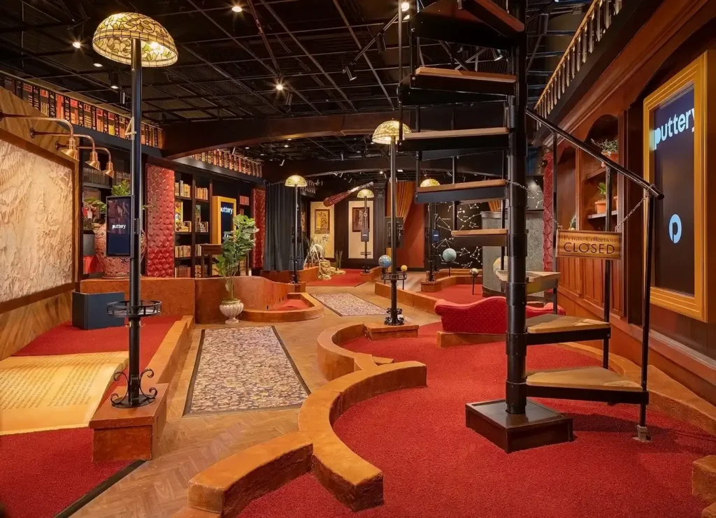 Indoor miniature golf setup with curved red carpet, wooden platforms, and a black metal spiral staircase under warm lights.