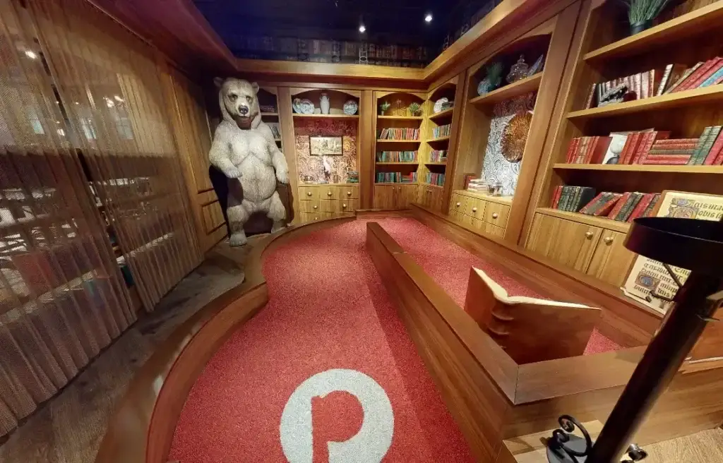 Library mini golf hole with a large bear figure, floor-to-ceiling bookshelves, and red putting turf at Puttery Chicago.