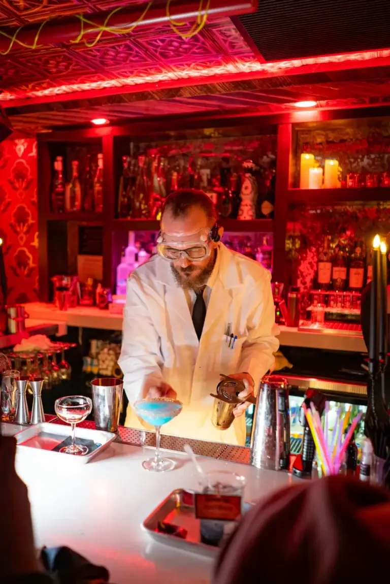 Bartender in a white coat and safety goggles mixing a blue-rimmed cocktail at a colorful bar with red lighting.