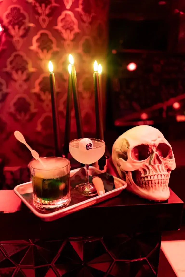 Halloween table with green cocktails, tall lit candles, and a skull prop under red lighting.
