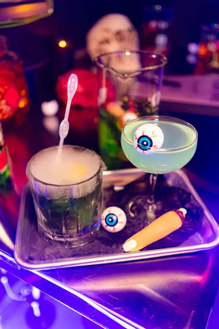 Colorful cocktails on a neon-lit bar, one with a blue eyeball garnish and surrounding decorations.