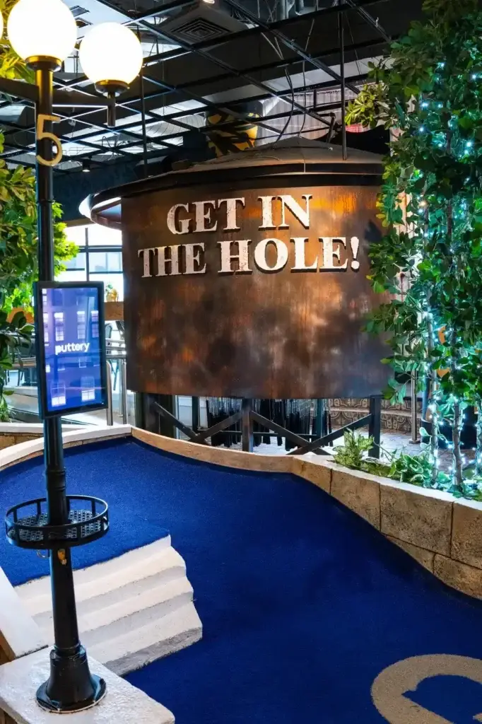 Blue turf mini golf hole with a large “Get In The Hole!” feature sign on the Rooftop course at Puttery Dallas.