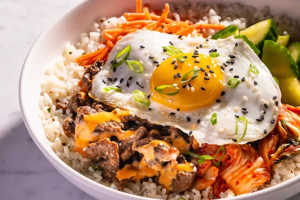 Korean Power Bowl at Puttery with marinated Korean beef, ginger rice, kimchi, marinated cucumbers and carrots, topped with a sunny side egg, green onions, sesame seeds, gochujang chili aioli and chili oil.