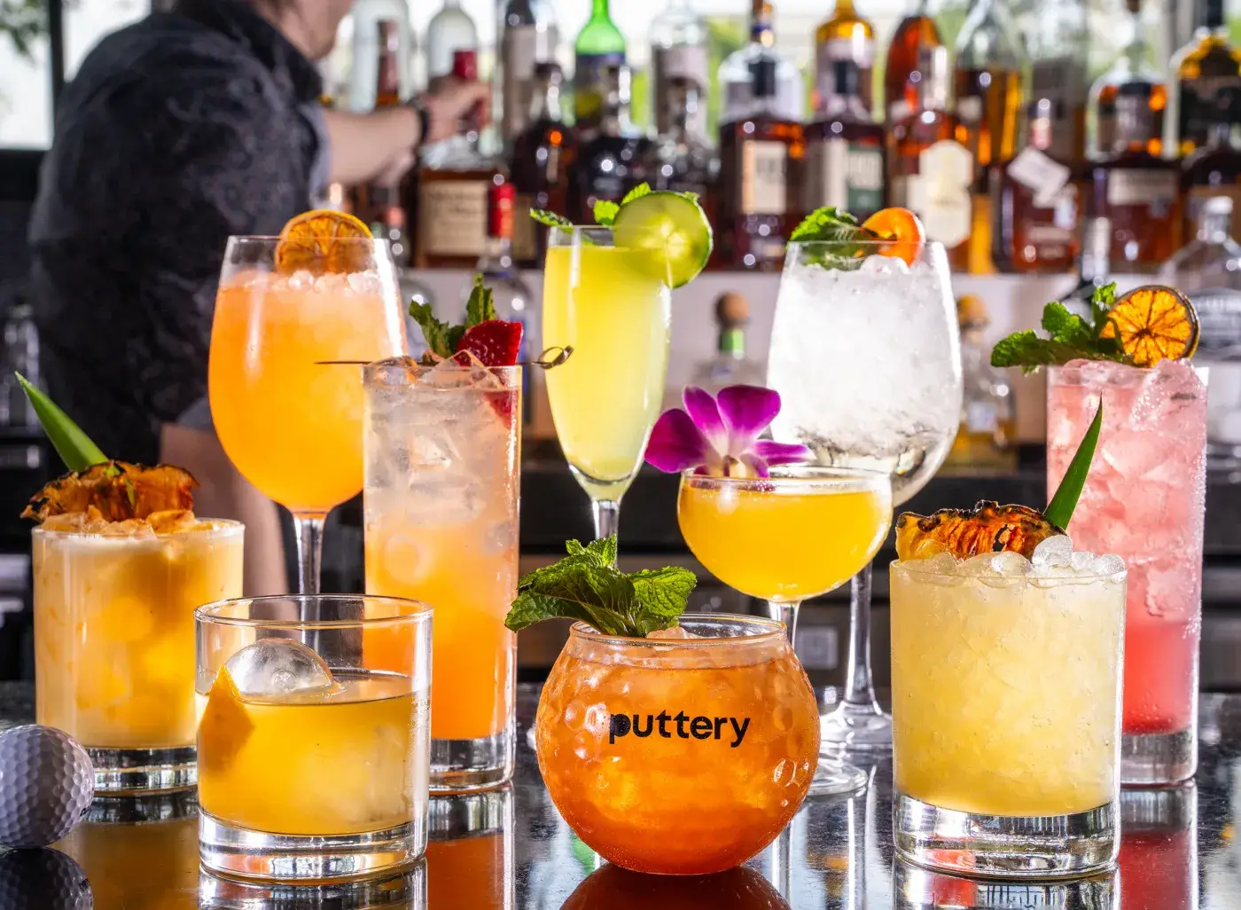 A lineup of colorful signature cocktails is arranged across a glossy bar top at Puttery. The drinks vary in glassware, color, and garnish, including citrus wheels, mint, pineapple, a purple orchid, strawberries, and crushed ice. Liquor bottles and a bartender appear blurred in the background, creating a lively bar setting.