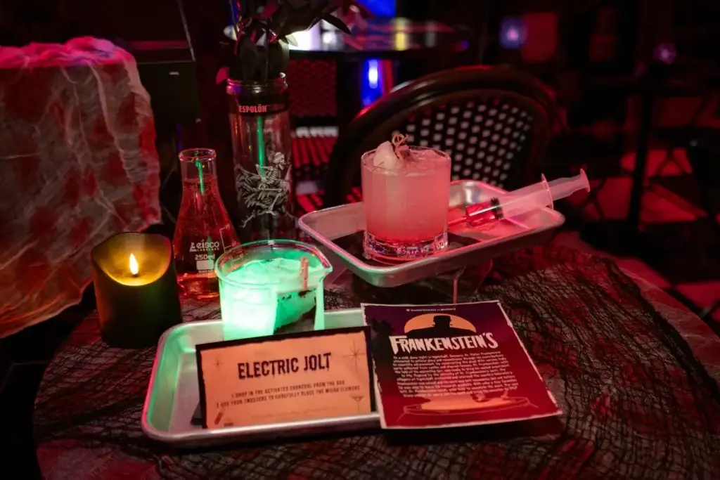 Interactive Frankenstein Cocktails at Puttery NYC - Puttery Close-up of glowing interactive Frankenstein cocktails with an Electric Jolt instruction card, syringe prop and lab glassware at Puttery NYC.