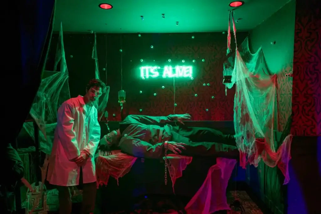 Costumed mad scientist beside a staged Frankenstein patient beneath a glowing green “IT’S ALIVE!” sign at Puttery NYC’s Rojo Room.