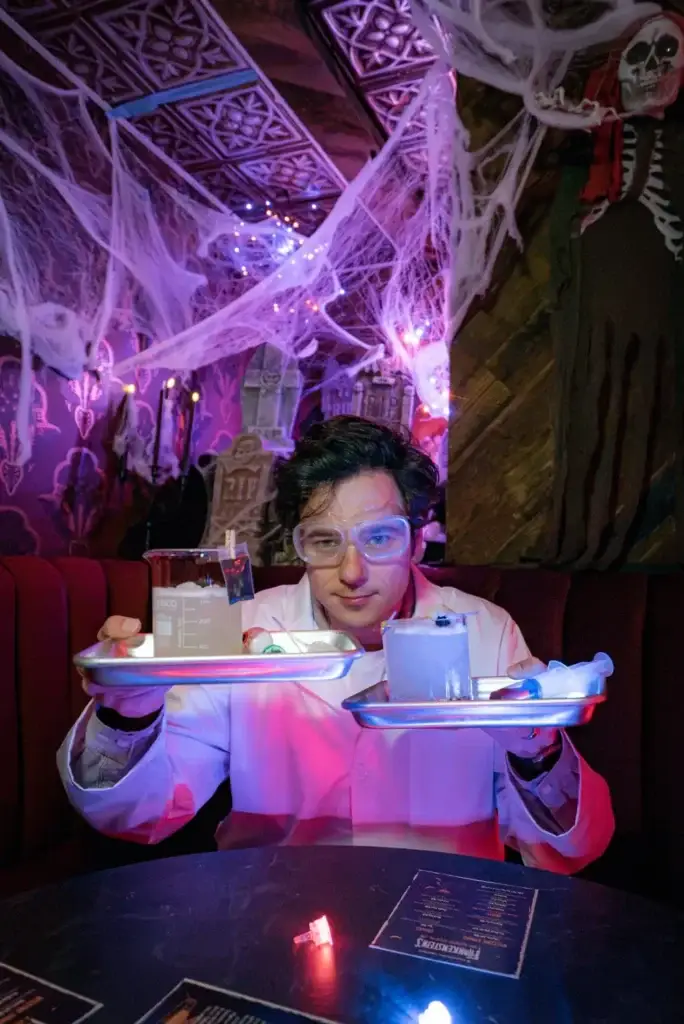 Mad Scientist Serving Interactive Cocktails at Puttery NYC - Puttery Mad-scientist bartender at Puttery NYC holding two metal surgical trays with glowing interactive cocktails, a beaker and syringe-style lab props.